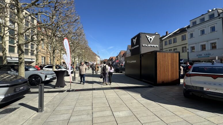 Eine sonnige Straßenszene während der Mobilitätstage zeigt Menschen, die an ausgestellten Autos und einem großen schwarzen CUPRA-Stand vorbeigehen. Laublose Bäume säumen den Gehweg, und auf beiden Seiten sind Gebäude mit Geschäften unter einem klaren blauen Himmel zu sehen.
