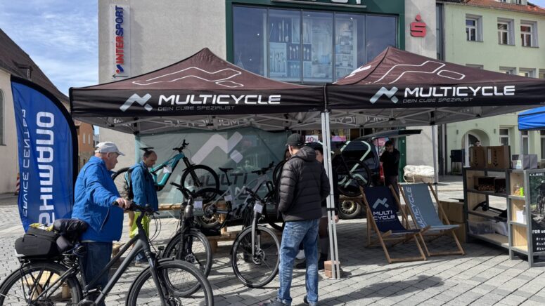 Menschen versammeln sich unter schwarzen Multicycle-Zelten bei einer Fahrradveranstaltung im Freien während der Mobilitätstage, um Fahrräder zu beobachten und zu testen. Die Szene spielt sich auf einem Stadtplatz mit Geschäften und Bannern im Hintergrund ab.