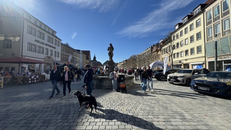 Ein belebter Platz mit Kopfsteinpflaster während der Mobilitätstage, mit spazierenden Menschen, Sitzgelegenheiten im Freien, einer Statue in der Mitte, geparkten Autos und einem schwarzen Hund im Vordergrund. Historische Gebäude säumen die sonnige Straße unter blauem Himmel.