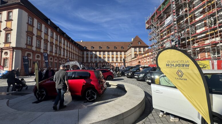 Auf einem sonnigen Platz unter freiem Himmel, umgeben von historischen Gebäuden, werden während der Mobilitätstage Autos ausgestellt; eine gelbe "Autohaus Weidlich Renault"-Fahne und ein rotes Auto stechen im Vordergrund hervor, ein Gebäude ist eingerüstet.