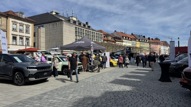 Während der Mobilitätstage, einer Autoausstellung im Freien, gehen Menschen zwischen geparkten Autos auf einer Kopfsteinpflasterstraße umher. Im Hintergrund sind Zelte, Gebäude mit bunten Fassaden und ein teilweise bewölkter Himmel zu sehen.