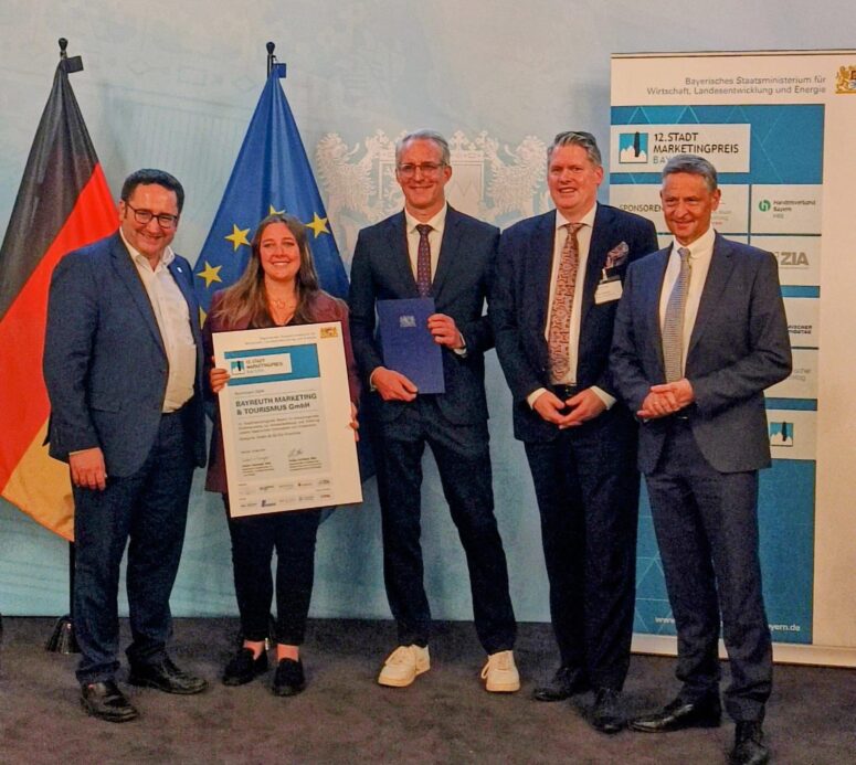 Fünf Menschen stehen in einem Raum vor deutschen und EU-Fahnen, lächeln und feiern ihre Erfolge. Eine Frau hält eine große Urkunde in der Hand, ein Mann neben ihr einen Ordner. Auf der rechten Seite steht ein Banner mit Logos und Text.
