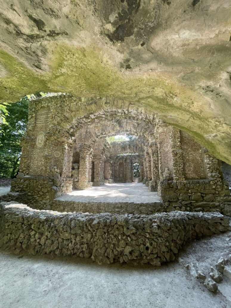 Ein steinerner Torbogen und Mauern aus unregelmäßigen Felsen bilden die Ruinen eines alten Bauwerks, das teilweise von einem überhängenden Felsen beschattet wird und durch das das Sonnenlicht in eine üppige, grüne Umgebung fällt.