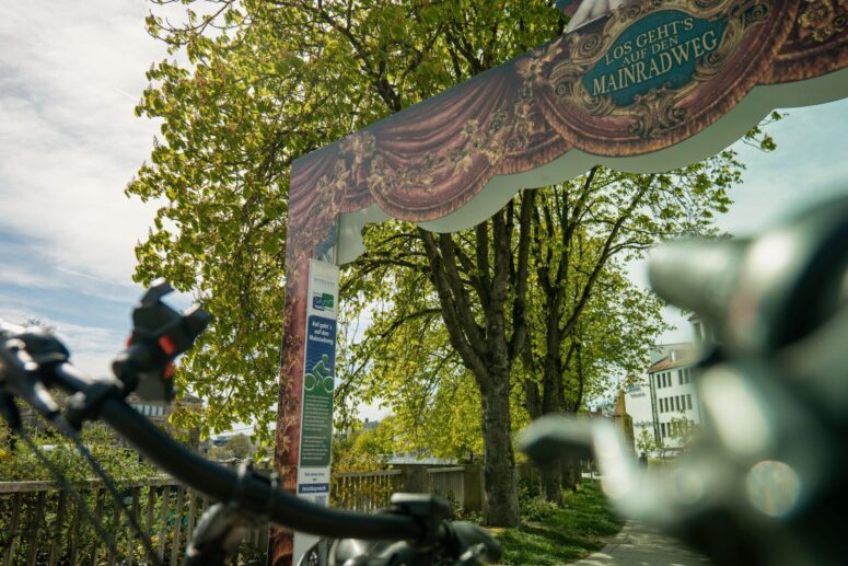Ein dekorativer Bogen mit der Aufschrift LOS GEHTS AUF DEN MAINRADWEG steht über einem von Bäumen gesäumten Radweg, mit einem Fahrradlenker im Vordergrund und Gebäuden, die an einem sonnigen Tag im Hintergrund zu sehen sind.