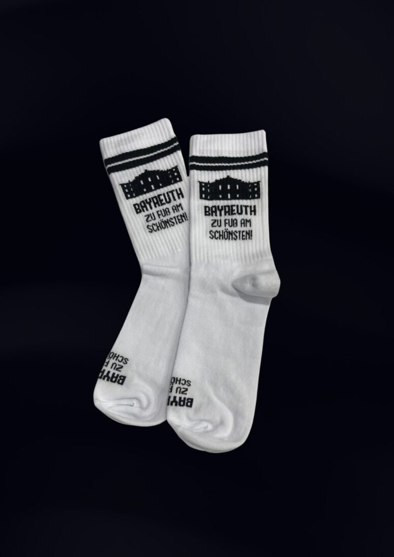 Zwei weiße Socken aus dem Bayreuth Shop mit schwarzem Text und Design, mit einem stilvollen schwarz-weiß gemusterten Band am oberen Rand. Unterhalb des Bandes ist "BAYREUTH ZU FUSS AM SCHÖNSTEN" aufgedruckt. Die Socken sind nebeneinander auf einem dunklen Hintergrund abgebildet.