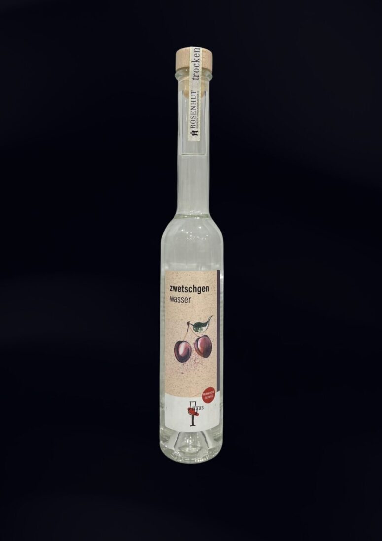 Eine Klarglasflasche mit hohem Hals und Korkenverschluss, beschriftet mit "Zwetschgenwasser". Das Etikett des Bayreuther Ladens zeigt zwei Pflaumen und einen kleinen Vogel vor einem schlichten schwarzen Hintergrund.