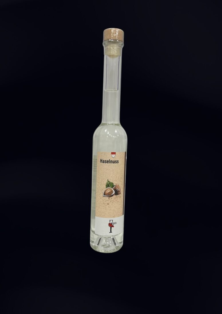 Eine hohe, klare Glasflasche mit einem Korkdeckel enthält eine farblose Flüssigkeit. Das Etikett trägt die Aufschrift Haselnuss und zeigt Haselnüsse mit grünen Blättern auf einem beigen Hintergrund. Erhältlich ausschließlich im Bayreuth Shop.