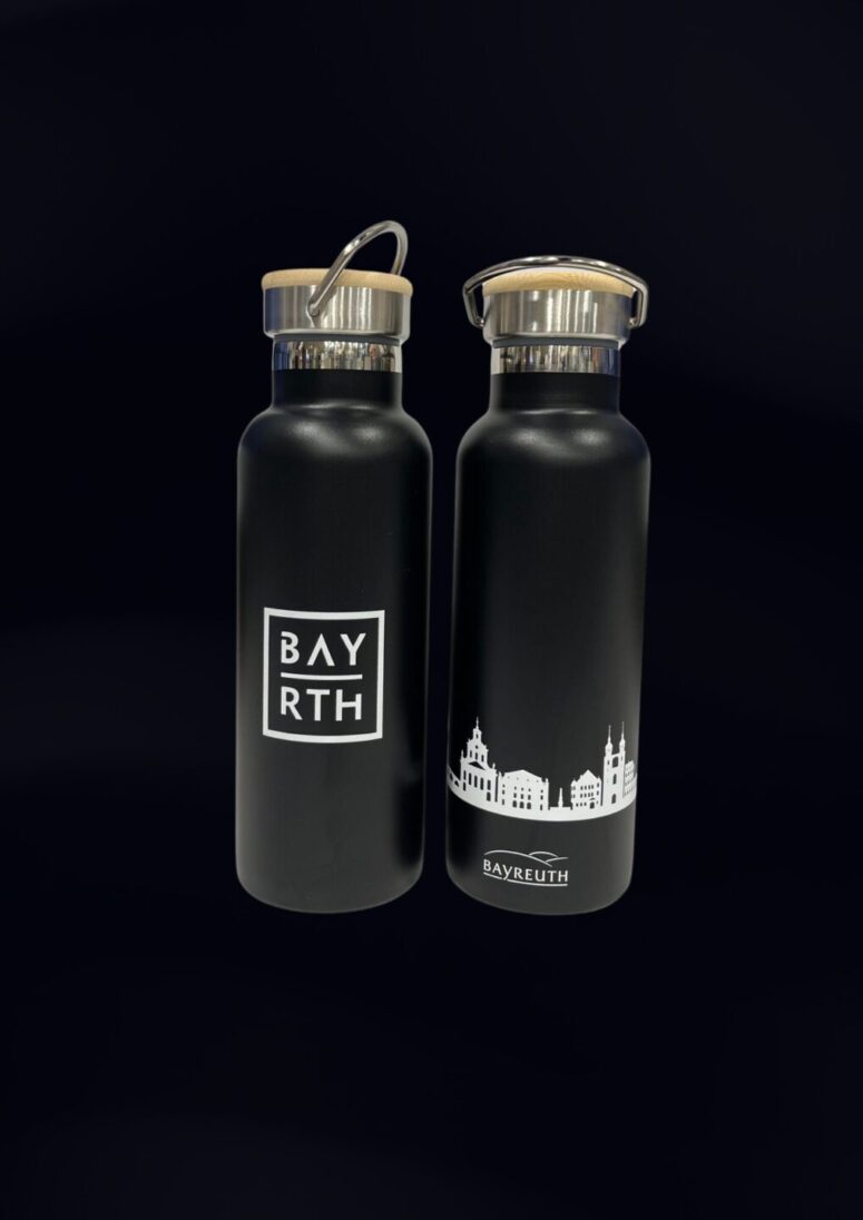 Zwei schwarze Wasserflaschen mit Holzdeckeln. Die Flasche auf der linken Seite zeigt ein weißes BAYRTH-Logo in einem Quadrat, während die Flasche auf der rechten Seite ein Stadtbild mit der Aufschrift BAYREUTH zeigt - jetzt erhältlich im Bayreuth Shop.