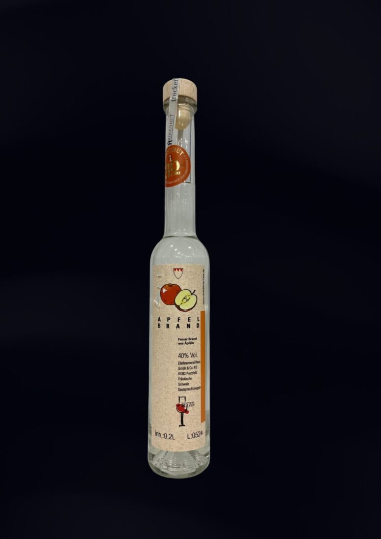 Eine hohe, schlanke Glasflasche mit Holzkappe, die eine klare Flüssigkeit enthält. Auf dem Etikett sind Äpfel und ein deutscher Text zu sehen, der ihn als Apfelbrand mit 40 Volumenprozent Alkohol bezeichnet. Erhältlich im Bayreuth Shop. Der Hintergrund ist schwarz.