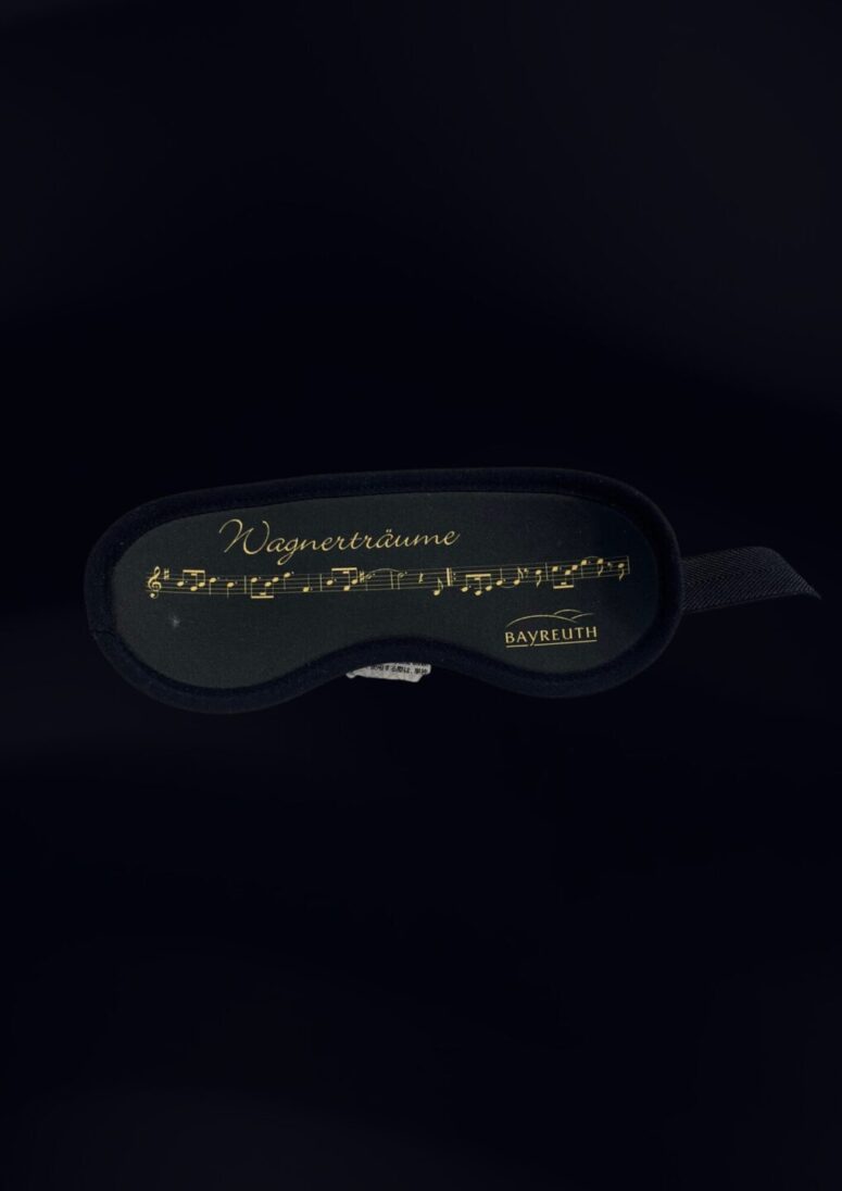 Eine schwarze Schlafmaske mit goldenen Noten, "Wagnerträume" und "BAYREUTH" auf der Vorderseite, erhältlich im Bayreuth Shop, fotografiert vor einem dunklen Hintergrund.