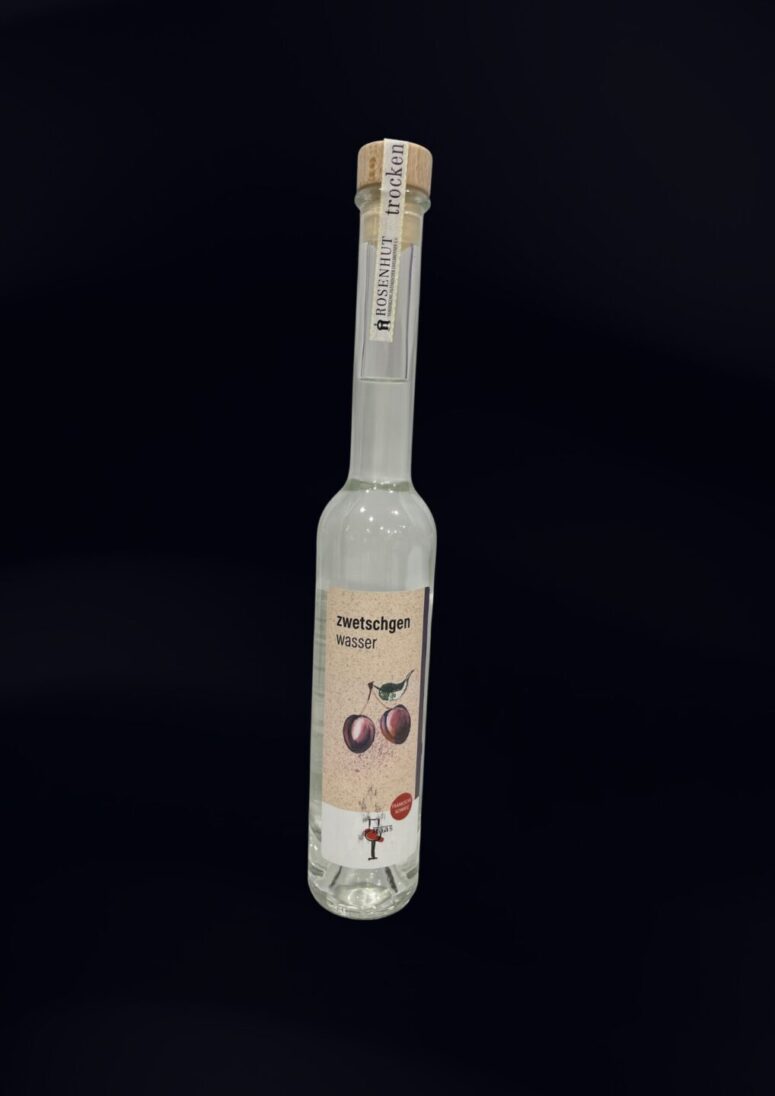 Eine klare Glasflasche mit Korken, die mit "zwetschgen wasser" beschriftet ist und auf deren Etikett zwei Pflaumen abgebildet sind, wird vor einem schlichten schwarzen Hintergrund präsentiert - exklusiv im Bayreuth Shop erhältlich.