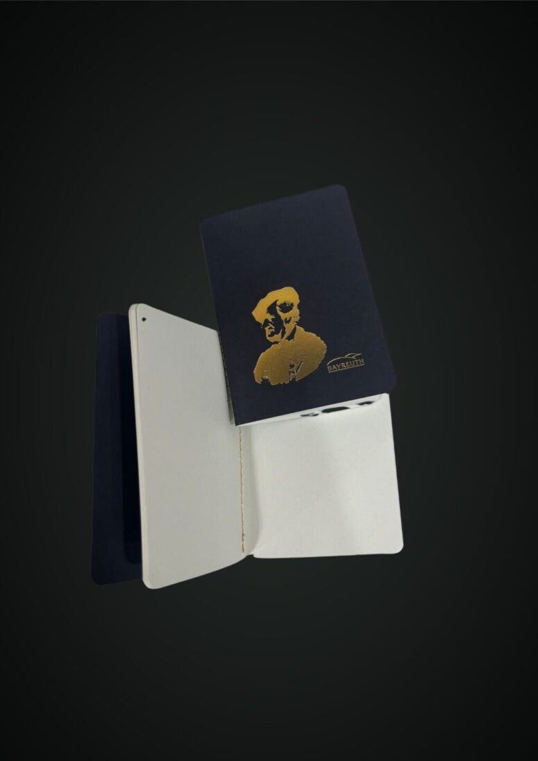 Zwei kleine Notizbücher, eines geschlossen und eines offen, mit dunklem Einband. Das geschlossene Notizbuch zeigt das in Gold geprägte Bild einer Person mit Brille und Hut sowie den Schriftzug CANVAS auf dem Einband - ab sofort im Bayreuth Shop erhältlich.
