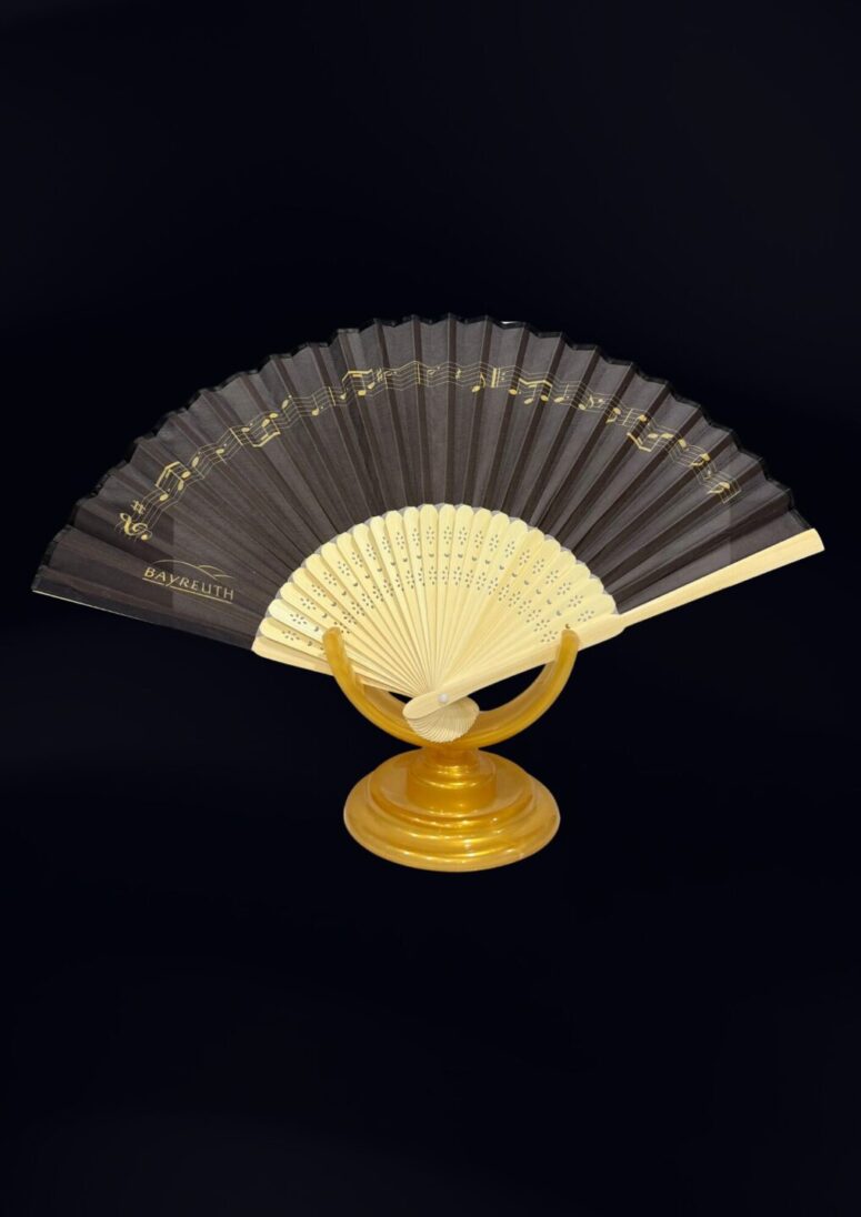 Der dekorative Handfächer aus dem Bayreuth Shop zeigt schwarze und cremefarbene Paneele, die mit Musiknoten verziert sind. Offen auf einem runden goldenen Ständer präsentiert, hebt er sich schön von einem soliden schwarzen Hintergrund ab.