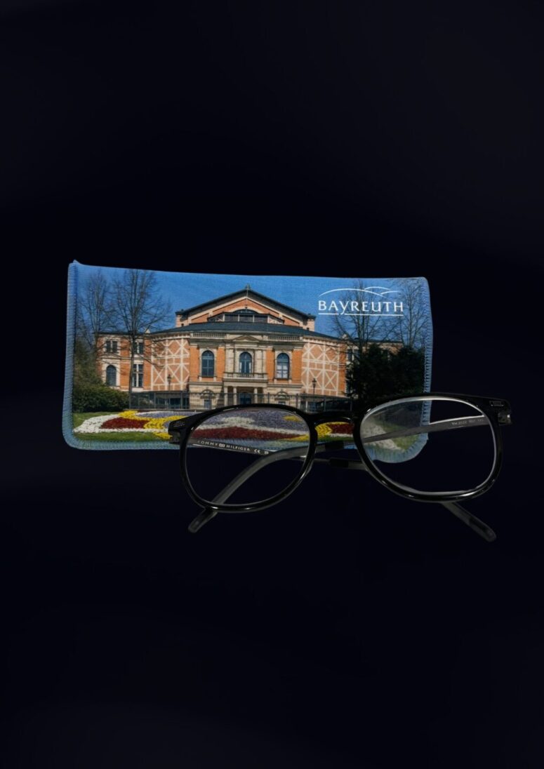 Eine schwarze Brille ruht vor einem Brillenetui mit dem Bayreuther Festspielhaus und dem Schriftzug Bayreuth" auf dunklem Hintergrund - ein elegantes Accessoire aus dem Bayreuth-Shop.