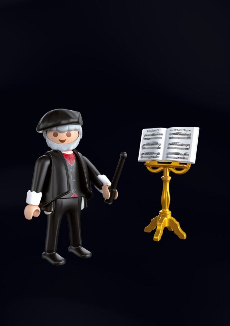 Eine Playmobil-Figur eines Dirigenten in schwarzer Kleidung und Hut hält einen Taktstock und steht neben einem goldenen Notenpult mit einem aufgeschlagenen Notenbuch - ein exklusives Fundstück aus dem Bayreuth-Shop - vor schwarzem Hintergrund.