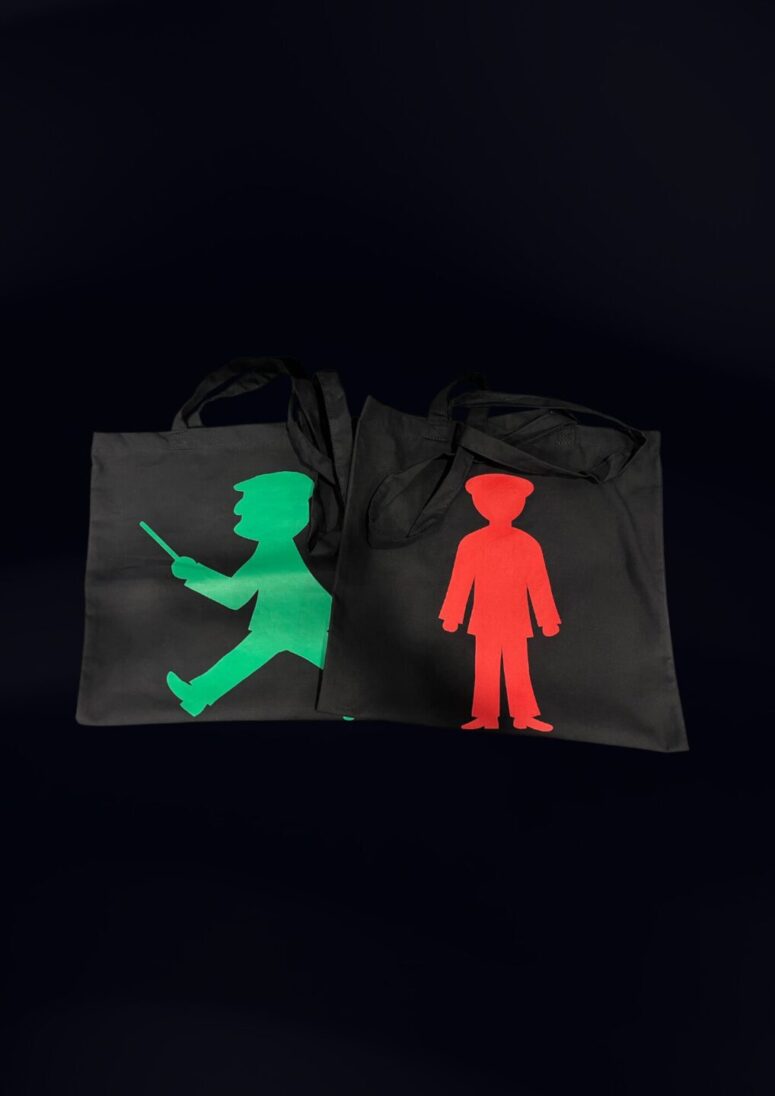 Zwei schwarze Tragetaschen auf dunklem Grund aus dem Bayreuth Shop; eine zeigt eine grüne, gehende Figur mit Hut und Stock, die andere eine rote, stehende Figur mit Hut, beide in Form von Fußgängerampel-Symbolen.