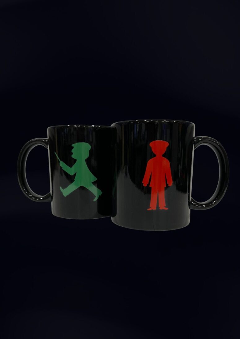 Zwei schwarze Tassen aus dem Bayreuth Shop vor dunklem Hintergrund; eine zeigt eine grüne gehende Figur und die andere eine rote stehende Figur, beide inspiriert von Fußgängerampelsymbolen.