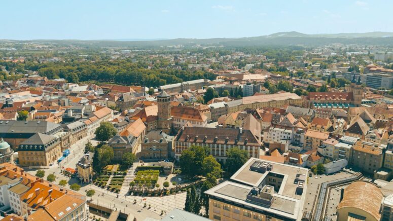 Luftaufnahme einer europäischen Stadt mit roten Ziegeldächern, historischen Gebäuden und von Bäumen gesäumten Straßen - perfekt für Pressefotos - vor der Kulisse grüner Hügel und eines klaren blauen Himmels.