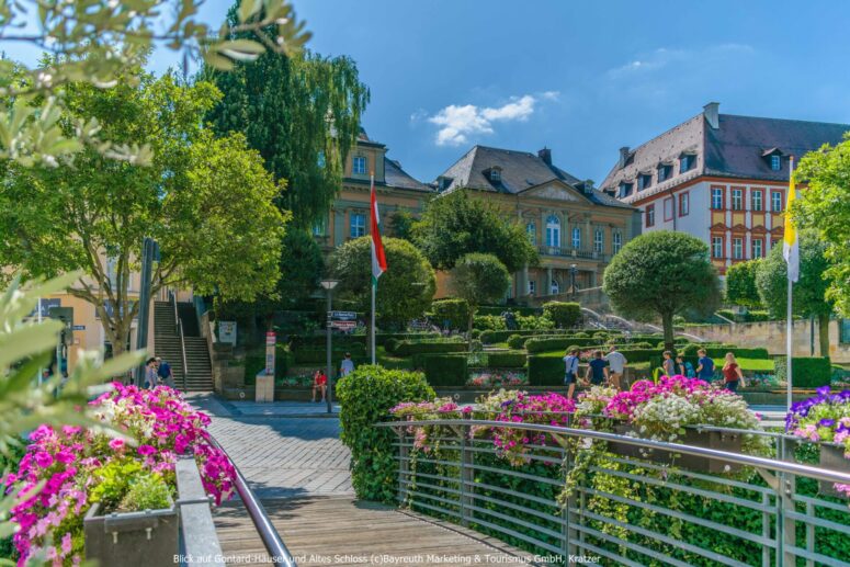 Ein sonniger Tag in einem Landschaftspark mit bunten Blumen, beschnittenen Büschen und Bäumen. Ein Spazierweg mit Geländer führt zu historischen Gebäuden mit verzierten Fassaden im Hintergrund. Diese CC0-Bilder-Szene zeigt Menschen, die spazieren gehen und sich entspannen.