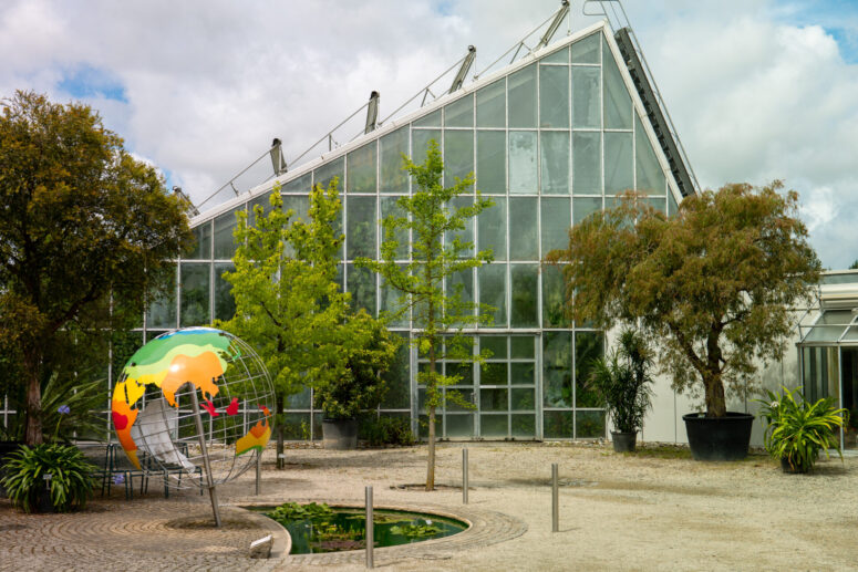 Hinter mehreren Bäumen und Pflanzen steht ein modernes Glasgewächshaus. Davor steht eine bunte Kugelskulptur neben einem kleinen runden Teich mit Seerosenblättern. Der Himmel ist in dieser CC0-Bilder-Szene teilweise bewölkt.