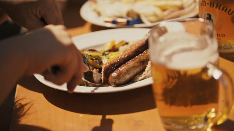 Eine Person schneidet in gegrillte Würstchen mit Gemüse auf einem weißen Teller, neben einem Glas Bier auf einem Holztisch in hellem Sonnenlicht. Im Hintergrund ist ein weiterer Teller mit Lebensmitteln zu sehen, alles in atemberaubender Fotografie im CC0-Bilder-Stil.