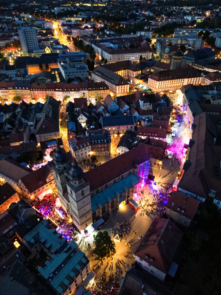Luftaufnahme einer nächtlichen Stadt mit hell erleuchteten Straßen und Gebäuden, Menschenmassen, die sich um eine große beleuchtete Kirche versammelt haben, und bunten CC0-Bildern, die eine festliche Atmosphäre schaffen.