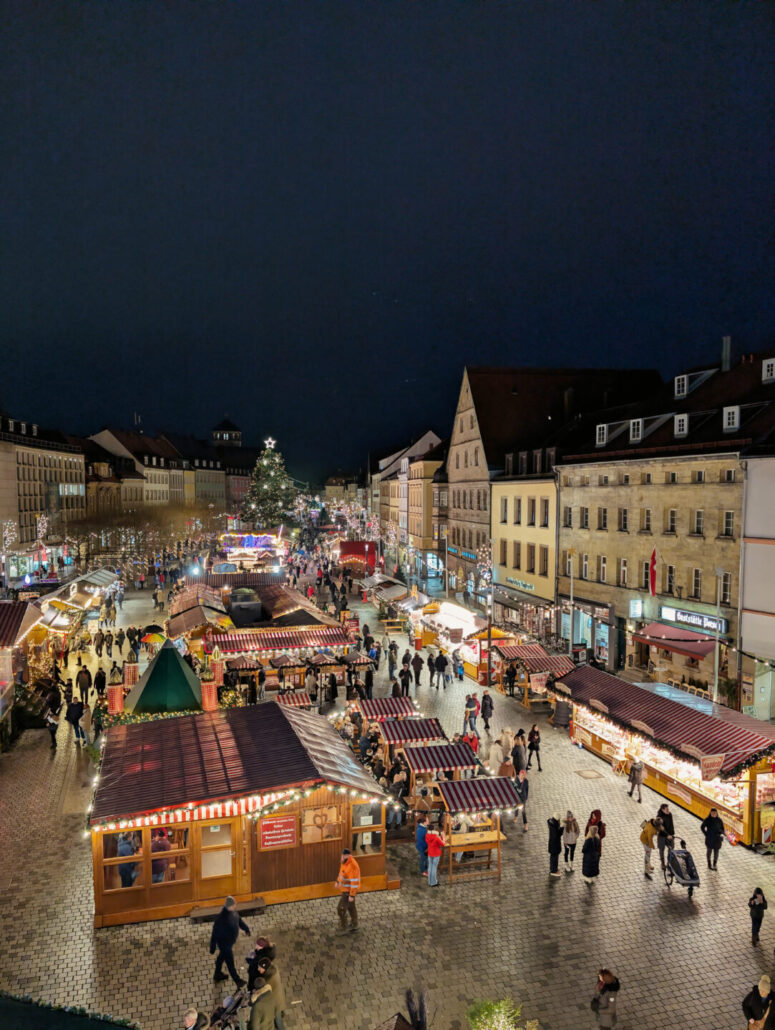 Ein festlicher nächtlicher Weihnachtsmarkt mit Menschen, die zwischen Verkaufsständen umhergehen, einem großen geschmückten Weihnachtsbaum im Hintergrund und historischen Gebäuden, die den beleuchteten Platz säumen - alles wunderschön in CC0-Bildern festgehalten.