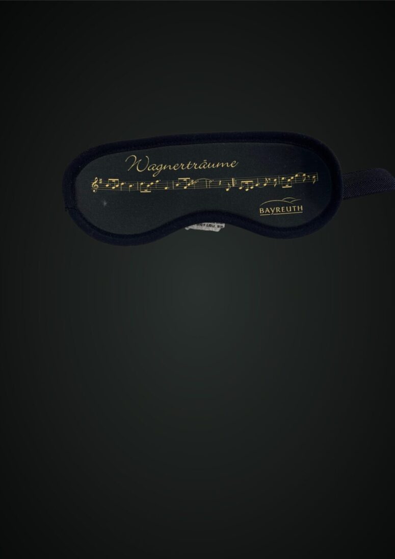 Eine schwarze Schlafmaske mit goldenen Noten und dem Schriftzug Wagnerträume darüber sowie Bayreuth unten rechts auf dunklem Hintergrund ist exklusiv im Bayreuth Shop erhältlich.