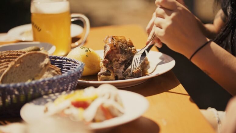 Eine Person schneidet in gebratenes Fleisch auf einem Teller, begleitet von einer Kartoffel und Soße. Auf dem sonnenbeschienenen Holztisch stehen ein Korb mit Brot, Salat, ein Glas Bier und andere Elemente, die typisch für einladende CC0-Bilder von Lebensmittelszenen sind.
