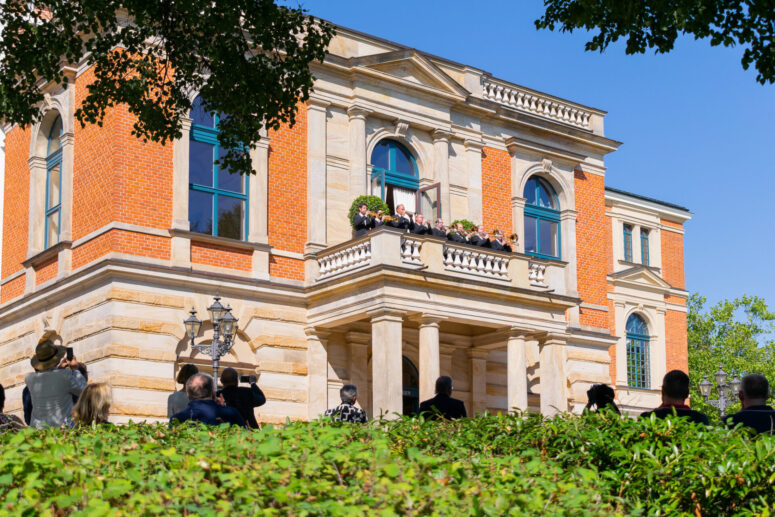 Eine Gruppe von Menschen steht auf dem Balkon eines prächtigen, historischen Gebäudes mit orangefarbenen Ziegeln und Steinmauern, aufgenommen in atemberaubenden CC0-Bildern, während andere vom Garten aus unter einem klaren, sonnigen Himmel zusehen und Fotos machen.