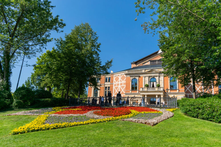 Ein großes Backsteingebäude mit einer dekorativen Fassade steht hinter einem farbenfrohen, herzförmigen Blumenbeet auf einem grünen Rasen, umgeben von hohen Bäumen unter einem klaren blauen Himmel - eine einladende Szene, die sich perfekt für CC0-Bilder-Projekte eignet.