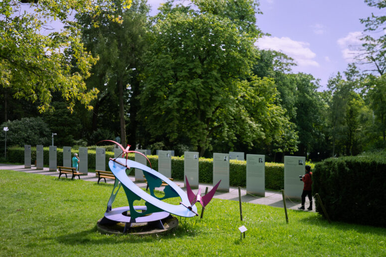 Bunte Metallskulptur auf einer Wiese in einem Park, mit CC0-Bildern auf einer Reihe von Informationstafeln und zwei Personen, die in der Nähe spazieren gehen. Üppig grüne Bäume und Laub füllen den Hintergrund unter einem teilweise bewölkten Himmel.