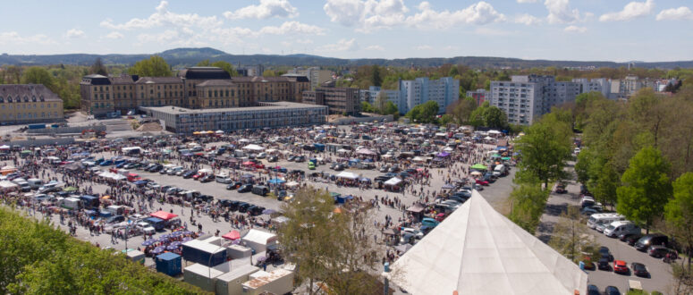 Ein großer Markt im Freien mit zahlreichen Ständen und Menschenmassen in einem Stadtgebiet mit Wohnhäusern und Bäumen. CC0-Bilder fangen ein weißes Zelt in der Nähe des Eingangs unter einem teilweise bewölkten Himmel ein.