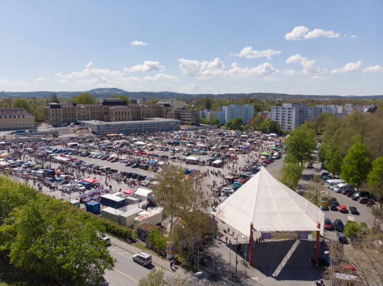 Ein großer Markt im Freien mit zahlreichen Ständen und Menschenmassen in einem Stadtgebiet mit Wohnhäusern und Bäumen. CC0-Bilder fangen ein weißes Zelt in der Nähe des Eingangs unter einem teilweise bewölkten Himmel ein.