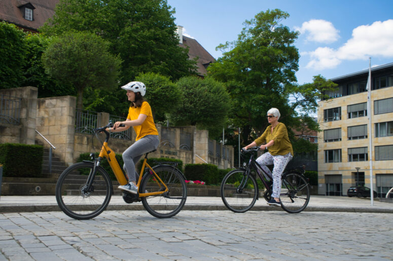 Zwei Personen, eine jüngere und eine ältere, fahren an einem sonnigen STADTRADELN-Tag auf einem gepflasterten Weg in einem Stadtgebiet mit Bäumen, Steintreppen und Gebäuden im Hintergrund Fahrrad.