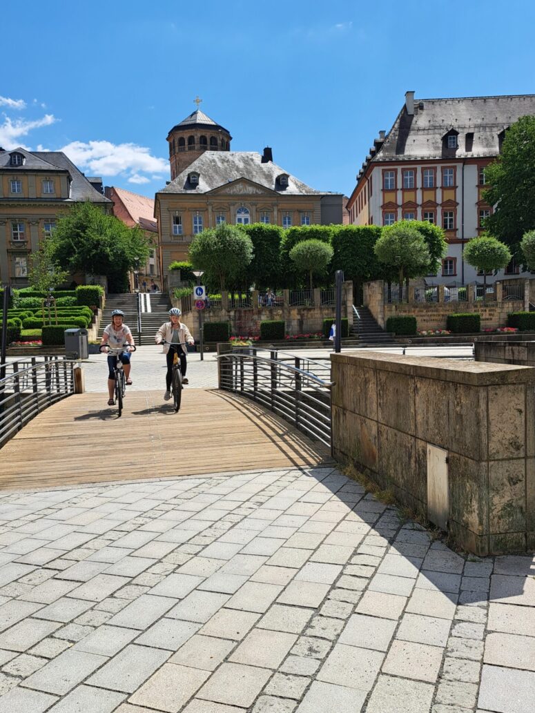 Zwei Personen fahren mit dem Fahrrad über eine Holzbrücke auf einem europäischen Stadtplatz während des STADTRADELNs, umgeben von historischen Gebäuden, beschnittenen Bäumen und einer Kuppelkirche unter einem strahlend blauen Himmel.