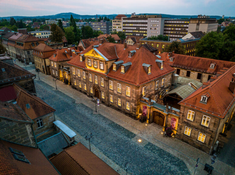 Luftaufnahme einer alten europäischen Stadt mit kopfsteingepflasterten Straßen und historischen Gebäuden, deren rote Ziegeldächer in der Abenddämmerung leuchten. In der Ferne sind moderne Gebäude und Grünflächen zu sehen. Das Foto ist ein CC0-Bilder.
