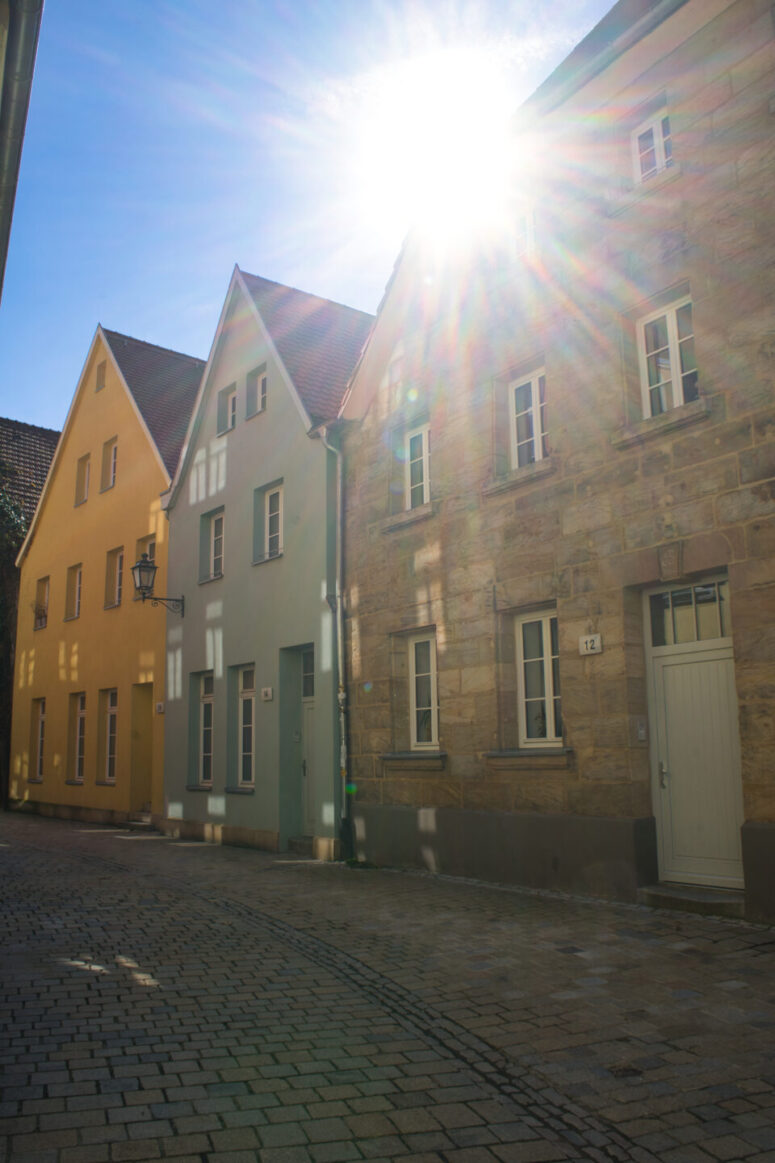Eine sonnenbeschienene Kopfsteinpflasterstraße mit drei benachbarten Häusern mit steilen Dächern in sanftem Gelb, Grün und Stein. Das Sonnenlicht scheint hell über die Dächer und wirft Schatten und Streulicht - perfekt für CC0-Bilder-Projekte.