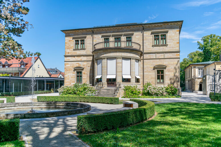 Ein historisches Steingebäude mit großen Fenstern und weißen Jalousien, umgeben von grünen Rasenflächen, einem runden Brunnen, gestutzten Hecken und Bäumen unter einem klaren blauen Himmel - eine ideale Kulisse für beeindruckende CC0-Bilder.