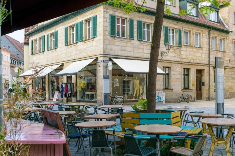 Ein Außencafé mit bunten Tischen und Stühlen befindet sich auf einer Kopfsteinpflasterstraße vor einem Bekleidungsgeschäft in einem historischen Steingebäude mit grünen Fensterläden. Diese ruhige, sonnige Straßenszene versprüht den Charme, den man oft in CC0-Bildersammlungen findet.