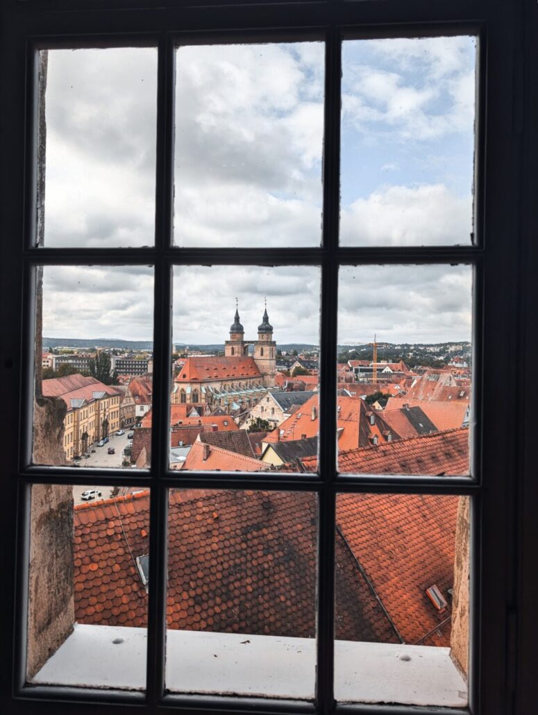 Blick durch ein Fenster auf eine europäische Stadtlandschaft mit roten Ziegeldächern, einer Kirche mit zwei Türmen und einem wolkenverhangenen Himmel darüber. Der Fensterrahmen unterteilt die Szene in Abschnitte. Perfekt für CC0-Bilder-Projekte, bei denen es auf authentischen städtischen Charme ankommt.