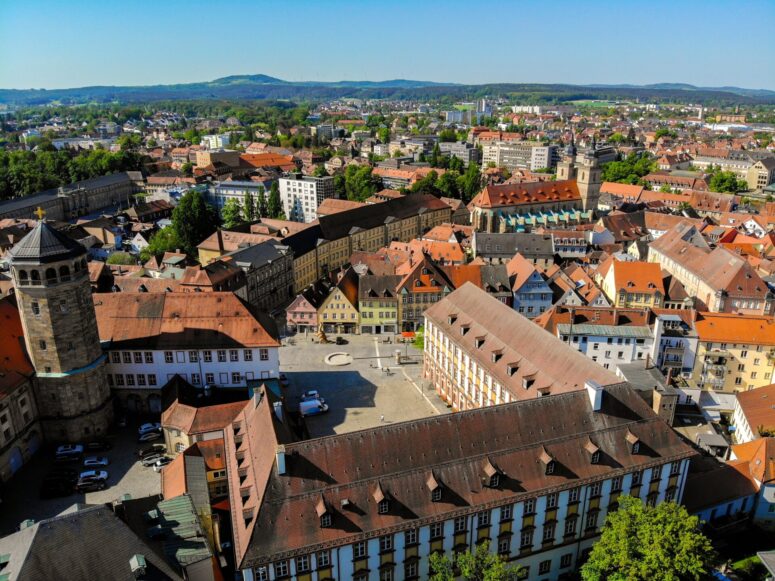 Luftaufnahme einer europäischen Stadt mit roten Ziegeldächern, historischen Gebäuden, einem zentralen Platz und einem runden Steinturm. Dahinter erstrecken sich üppig grüne Hügel, alles in lebendiger Klarheit eingefangen. Perfekte CC0-Bilder für Ihre Projekte unter einem klaren blauen Himmel.
