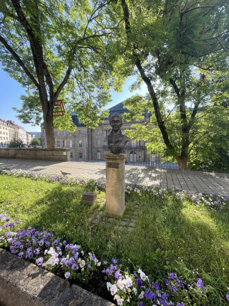 Eine Bronzebüste auf einem Steinsockel steht umgeben von violetten und weißen Blumen in einem sonnenbeschienenen Park, mit Bäumen und historischen Gebäuden im Hintergrund. Dieses CC0-Bilder fängt eine heitere, zeitlose Szene ein, die sich perfekt für kreative Projekte eignet.