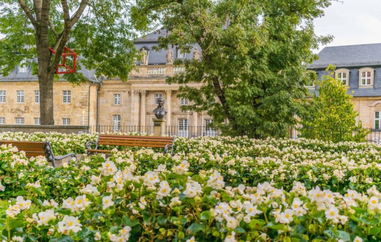 Ein Garten voller blühender weißer Blumen und Bänke, mit einer Statue und einem historischen Steingebäude im Hintergrund, teilweise beschattet von großen grünen Bäumen - perfekt für die Aufnahme schöner CC0-Bilder.