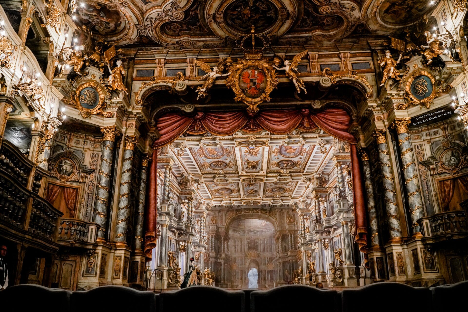 Opulenter Opernhaus-Innenraum mit kunstvollen Goldverzierungen, roten Vorhängen, aufwendigen Fresken, Kronleuchtern und detaillierten Decken. Die Bühne und die Balkone sind sichtbar und zeigen die großartige Architektur und das üppige Design.