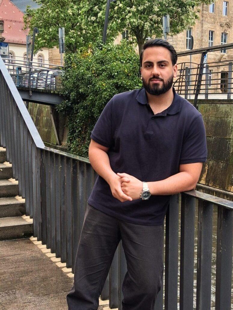 Ein bärtiger Mann in dunklem Polohemd und Hose lehnt lässig an einem Metallgeländer an einem Kanal, im Hintergrund sind Bäume und Steinhäuser zu sehen.