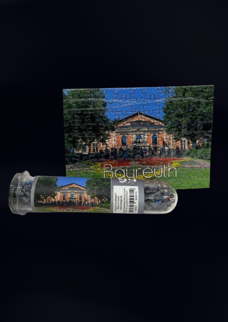 Ein fertiges Puzzle, das ein von Bäumen umgebenes Theatergebäude zeigt, steht aufrecht, davor liegt der Puzzleteil-Container des Bayreuth Shops mit der Beschriftung Bayreuth.