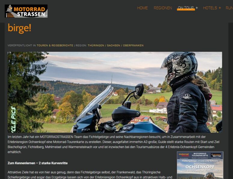 Ein Motorradfahrer mit Helm und Jacke sitzt auf einem Motorrad und genießt den Blick auf ein malerisches Dorf mit roten Dächern und herbstlichen Bäumen - die perfekte Inspiration für alle, die sich für Motorradfahren in Deutschland begeistern.