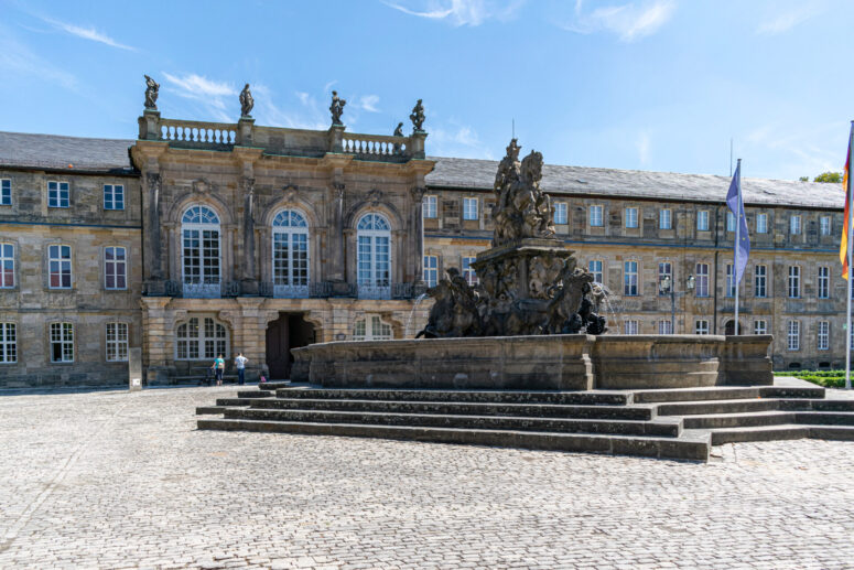 Ein großer Steinbrunnen mit Statuen steht vor einem historischen Gebäude mit gewölbten Fenstern, Säulen und Skulpturen auf dem Dach, unter einem klaren blauen Himmel. CC0-Bilder zeigen den lebhaften Platz mit Kopfsteinpflaster und flatternden Fahnen in allen Einzelheiten.