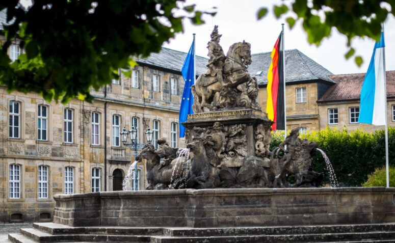 Ein Steinbrunnen mit kunstvollen Figuren und Pferden steht vor einem historischen Gebäude, das mit europäischen, deutschen und bayerischen Fahnen geschmückt ist. Baumzweige umrahmen die Szene darüber. Dieses CC0-Bilder Bild fängt den klassischen europäischen Charme ein.